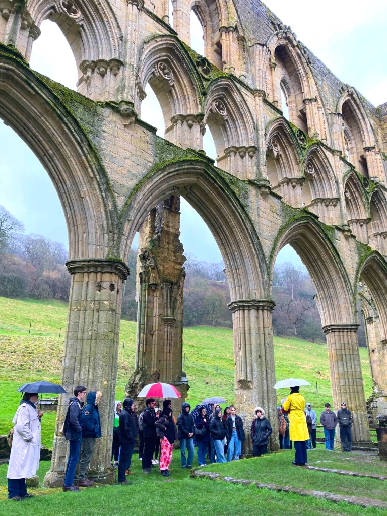 Group of students, some with umbrellas, listening to a talk in the arches of the Rievaulx Abbey church